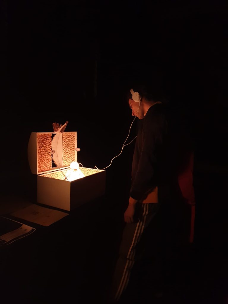 Fruizione della mostra: ragazzo che osserva lo scrigno aperto nel buio ascoltando il brano relativo tramite gli auricolari collegati allo scrigno